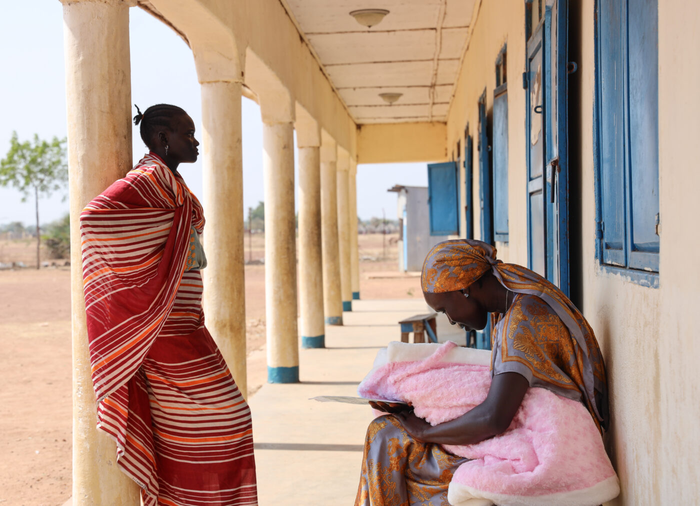 Two South Sudanese women wait outside a health facility. One of them holds a newborn